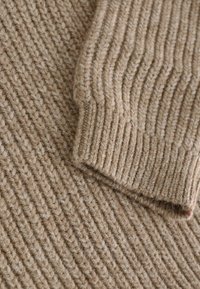 Beiger gestrickter Pulloverstoff mit vertikalen Rippen, der eine strukturierte, weiche Optik und einen gefalteten Ärmelabschluss aufweist.