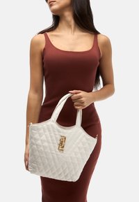 Sac fourre-tout matelassé blanc avec poignées doubles, détail de logo doré et forme structurée, doté d'une fermeture éclair et d'une texture lisse.