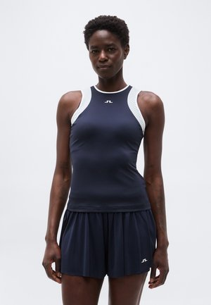 Atletska ženska, oblečena v temnoplavo brezrokavnik z belim trakcem in ujemajoče nabrane kratke hlače, stoji pred enobarvnim ozadjem.