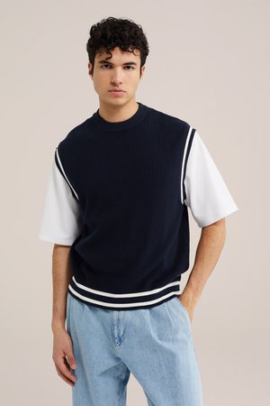 Giovane uomo con capelli ricci scuri che indossa un maglione senza maniche blu navy su una camicia bianca a maniche corte e jeans azzurri, in piedi con una mano in tasca.