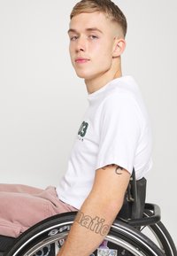 Camiseta corta de manga blanca con texto verde, pantalones rosas claros y una silla de ruedas moderna con un borde de rueda estampado y marco negro.