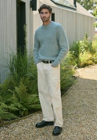 Lys blåt strikket sweater, creme bukser, sort bælte og sko. Baggrunden viser grønne planter og en grussti.