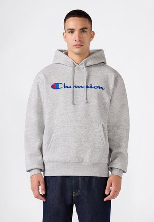 Junger Mann trägt einen grauen Champion-Hoodie mit Fronttasche und dunkelblauer Jeans, steht vor einem einfarbigen weißen Hintergrund.