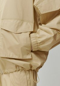Beige Windbreaker mit elastischen Bündchen, zwei seitlichen Taschen und einem Kordelzugsaum. Glatte Textur mit matter Oberfläche.