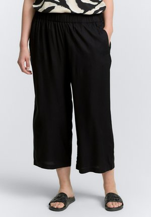 Pantalones cortos negros con una cintura elástica, fabricados en un tejido suave y con bolsillos laterales, combinados con sandalias negras tipo slide con un diseño trenzado.