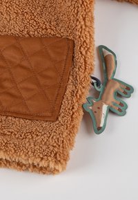 Tessuto in pile marrone morbido con una tasca trapuntata marrone e un piccolo giocattolo di stoffa a forma di volpe attaccato, con contorno verde.