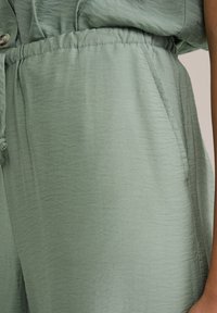 Ljusgrön, strukturerad jumpsuit i tyg med elastisk midja, sidofickor och knappdetaljer högst upp i en avslappnad design.