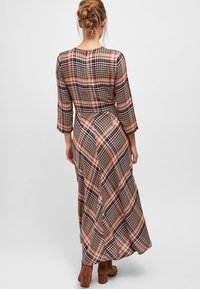 Femme aux cheveux blonds coiffés en chignon lâche, portant une longue robe à carreaux rose, marron et noire et des bottes à talons bruns, de dos sur un fond uni.