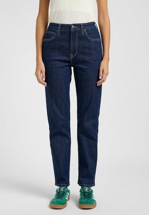 Femme portant un jean bleu foncé taille haute jambe droite avec des baskets vertes et blanches sur un fond gris clair.