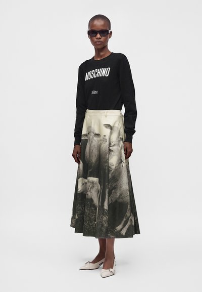 Femme portant un sweat-shirt noir Moschino, une jupe beige et noire avec un imprimé moutons, des chaussures blanches pointues et des lunettes de soleil noires, debout sur fond blanc.
