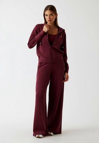 Weinroter Trainingsanzug aus weichem Stoff mit Kapuzenjacke und weit geschnittenen Hosen. Das Design umfasst gerippte Bündchen und einen eng anliegenden Tanktop darunter.