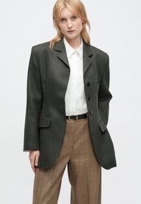 Blonde vrouw die een oversized donkergrijze gepinde blazer, een witte overhemd, en een bruine ruitbroek met zwarte riem draagt, staande tegen een witte achtergrond.