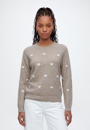 Donna con lunghe trecce, che indossa un maglione beige con motivi a cuore bianchi e pantaloni bianchi, in piedi su uno sfondo semplice.