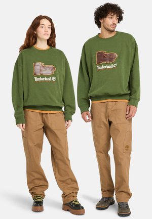 Green sweatshirt featuring a boot graphic and "Timberland Established 1973" lettering. Paired with tan cargo pants. Black and brown shoes.
