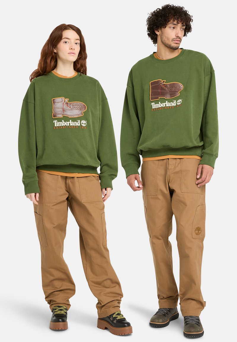 Green sweatshirt featuring a boot graphic and "Timberland Established 1973" lettering. Paired with tan cargo pants. Black and brown shoes.