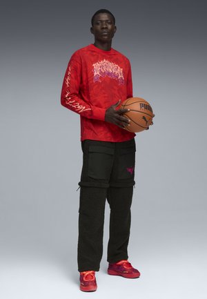 Jeune homme tenant un ballon de basket, portant un t-shirt à manches longues rouge avec un motif graphique, un pantalon noir texturé avec des poches et des baskets rouges sur un fond gris.