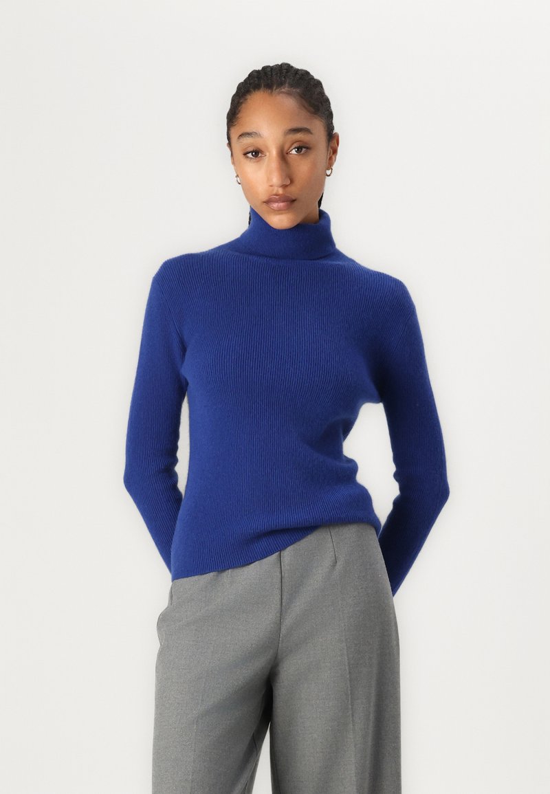 Jersey de cuello alto azul en textura acanalada con mangas largas, combinado con pantalones gris claro. Diseño simple y ajustado sin adornos.