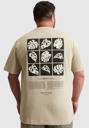 Beige T-Shirt mit einem schwarzen Grafikdruck von abstrakten Pflanzenblättern auf der Rückseite, darunter in Weißer Schrift. Kurzärmeliger Schnitt und lockere Passform.