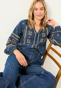 Marinebluse mit gestickten Blumenmustern, kombiniert mit hochgeschnittenen blauen Denim-Hosen; Modell sitzt auf einem Holzstuhl.