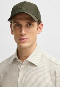 Olive green cap with "BOSS" logo, structured shape, smooth fabric. Worn with a striped beige and white collared shirt.