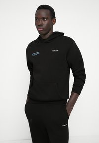 Czarna bluza z kapturem z kieszenią z przodu, ozdobiona białym i niebieskim tekstem. Dopasowana do czarnych spodni, obie mają luźny krój i ściągacze.