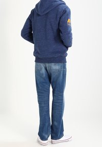 Sudadera navy con cordón y logo en la manga, combinada con jeans azules desgastados y zapatillas blancas. Enfocándose en un ajuste casual y cómodo.