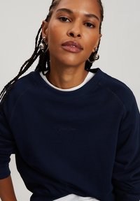 Granatowa bluza z okrągłym dekoltem, ściągaczami na rękawach i subtelnym wytłoczonym logo. Miękka tkanina w luźnym kroju z rękawami raglanowymi.