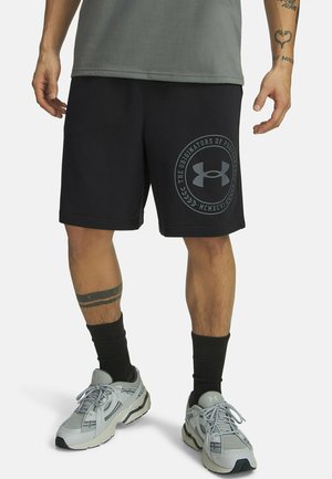 Mężczyzna noszący czarne spodenki Under Armour z logo, czarne skarpetki, szare buty do biegania i widoczne tatuaże na ramionach i jednej nodze.
