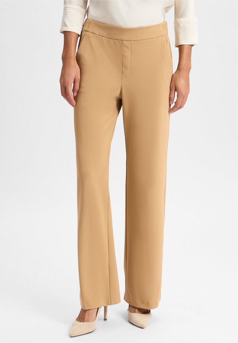 Pantalon beige à jambes larges en tissu lisse, doté d'une taille haute et de coutures discrètes, associé à des chaussures à talons neutres.