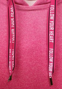Pink hoodie with textured fabric, featuring contrasting drawstrings printed with "FOLLOW YOUR HEART" in bold white text.