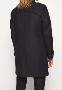 Cappotto di lana nera con spalle strutturate, polsini con bottoni e una vestibilità su misura. Presenta una texture liscia e linee pulite.