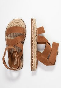 Bruna mockasin-sandaler i ruggat läder med flätade jute-sulor, breda korsade remmar och spänne vid ankeln, visade från ovan- och sidovy.