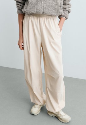 Personne portant un pantalon ample couleur crème, des baskets crème et blanches, et une veste polaire grise avec les mains dans les poches.