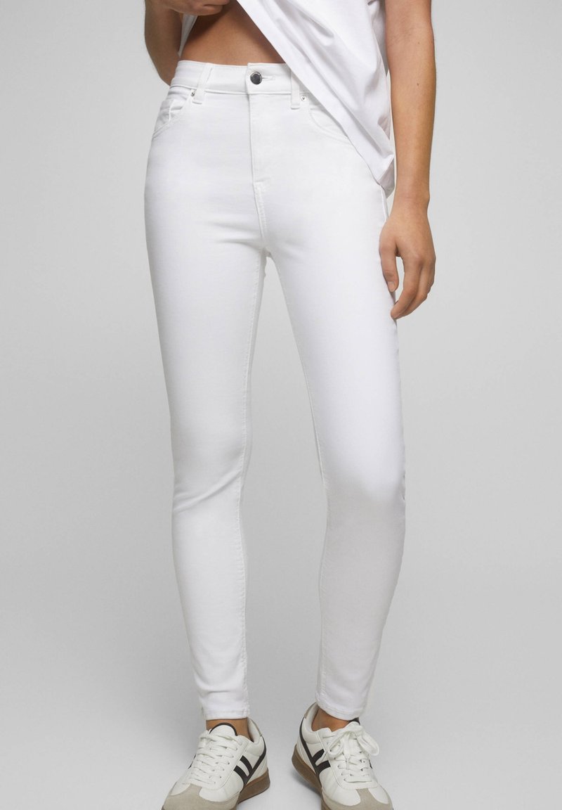 Femme portant un jean skinny blanc taille haute et des baskets blanches avec des rayures noires, levant son haut pour révéler une partie de son ventre.