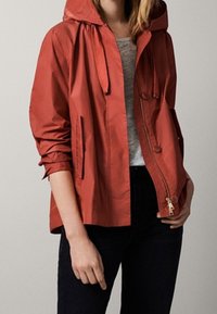 Kvinna som bär en rostorange huva jacka över en grå skjorta och svarta byxor, med jackan delvis öppen och händerna avslappnade.