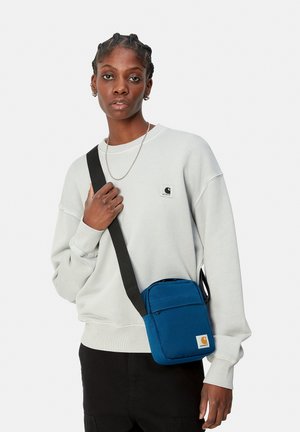 Bolso cruzado azul con una tela texturizada, que cuenta con un bolsillo frontal con cremallera y un parche con el logo. La correa ajustable es negra.