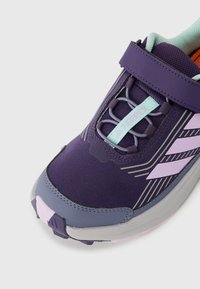 Chaussure de sport violette avec un dessus en maille texturée, une sangle réglable, un bout arrondi et des accents en bleu clair et lavande.