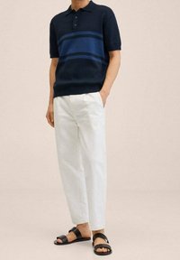 Hombre con polo navy a rayas azules, pantalones blancos y sandalias negras de doble correa, de pie con una mano en el bolsillo frente a un fondo liso.