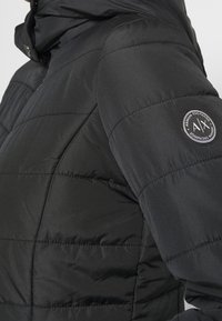 Chaqueta acolchada negra con un alto cuello, con costuras horizontales y un parche de logo circular en el hombro. Textura suave.
