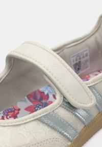 Chaussure Mary Jane beige avec une bride à velcro. Comprend une semelle intérieure à motif floral, des rayures argentées et un matériau supérieur texturé.