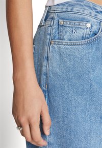 Jean en denim bleu clair à taille haute, avec un petit logo CK sur la poche arrière et des détails en métal visibles.