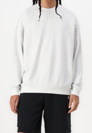 Personne portant un sweat-shirt Nike gris clair à manches longues et un short Nike noir avec poches latérales, debout devant un fond blanc.
