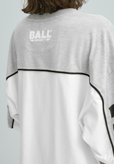 Sivá a biela nadmerná tričko s čiernymi akcentami. Na zadnej strane sa nachádza slovo "BALL" v bielej farbe a má uvoľnený strih s spadnutými plecami.