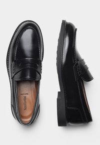 Mocassins en cuir noir avec une finition brillante, dotés d'une bande décorative et de détails de couture, ainsi qu'une semelle en caoutchouc plate.