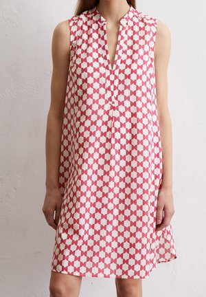 Vestido blanco sin mangas con patrón geométrico de estrellas rojas, cuello en V y abertura lateral, llevado por una persona de pie frente a una pared clara.