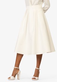 Jupe blanche, longueur genoux, avec une taille ajustée, un design plissé et une texture lisse. Associée à des sandales blanches à talons hauts avec des brides de cheville.