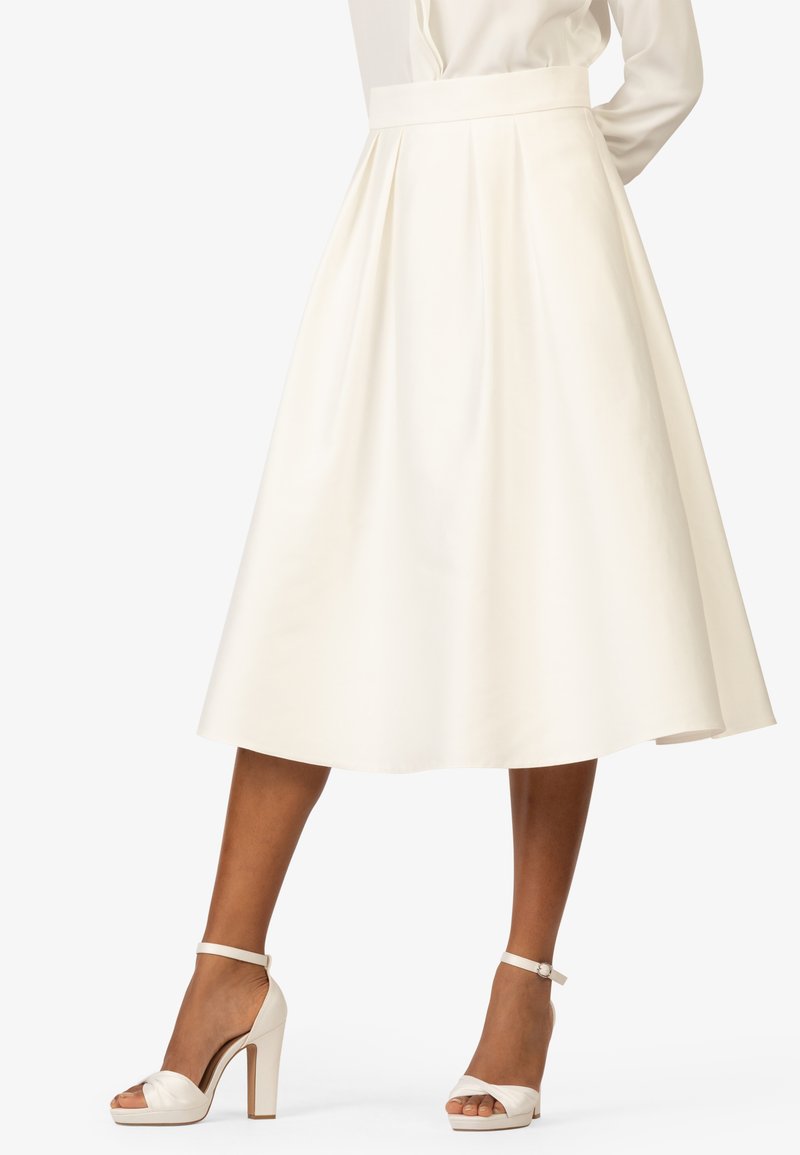 Jupe blanche, longueur genoux, avec une taille ajustée, un design plissé et une texture lisse. Associée à des sandales blanches à talons hauts avec des brides de cheville.