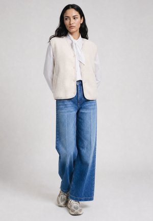 Woman wearing a white blouse with a bow, cream buttoned vest, wide-leg blue jeans, and gray sneakers standing against a plain background.