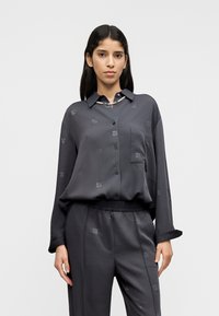 Σκούρα γκρι σατέν μπλούζα με κουμπιά στη μπροστινή όψη, γιακά και τσέπη, με διακριτικά μοτίβα λογότυπου. Συνδυάζεται με παντελόνι από σατέν στον ίδιο χρωματικό τόνο.