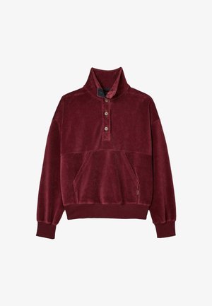 Burgunderfarbener Cordpullover mit hohem Kragen, Knopfdetails und einer Fronttasche, kombiniert mit braunen, karierten Schlaghosen und beigen Sneakers.